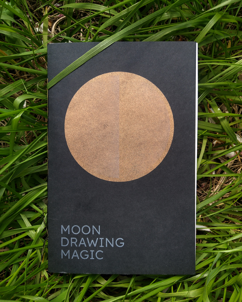 A book lays closed on a bed of fragrant sweet grass. The cover reads: Moon Drawing Magic.