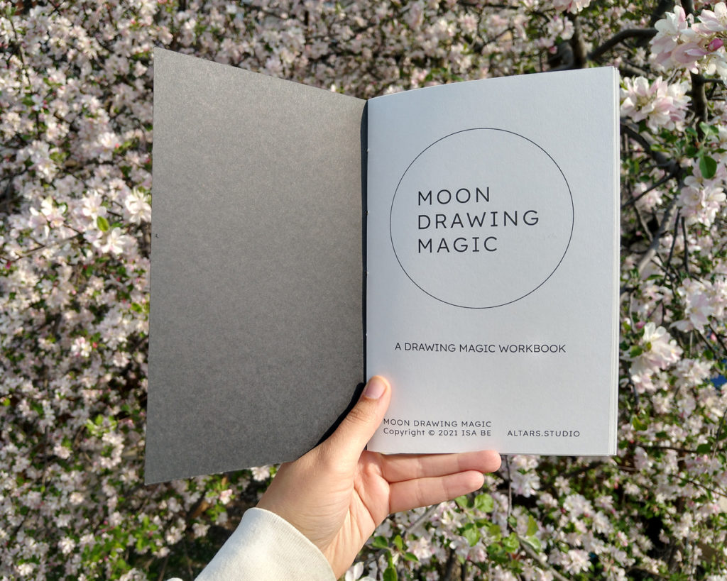 A hand holds a book open to the title page, which reads: Moon Drawing Magic, A Drawing Magic Workbook. Copyright 2021 Isa Be, Altars Studio.