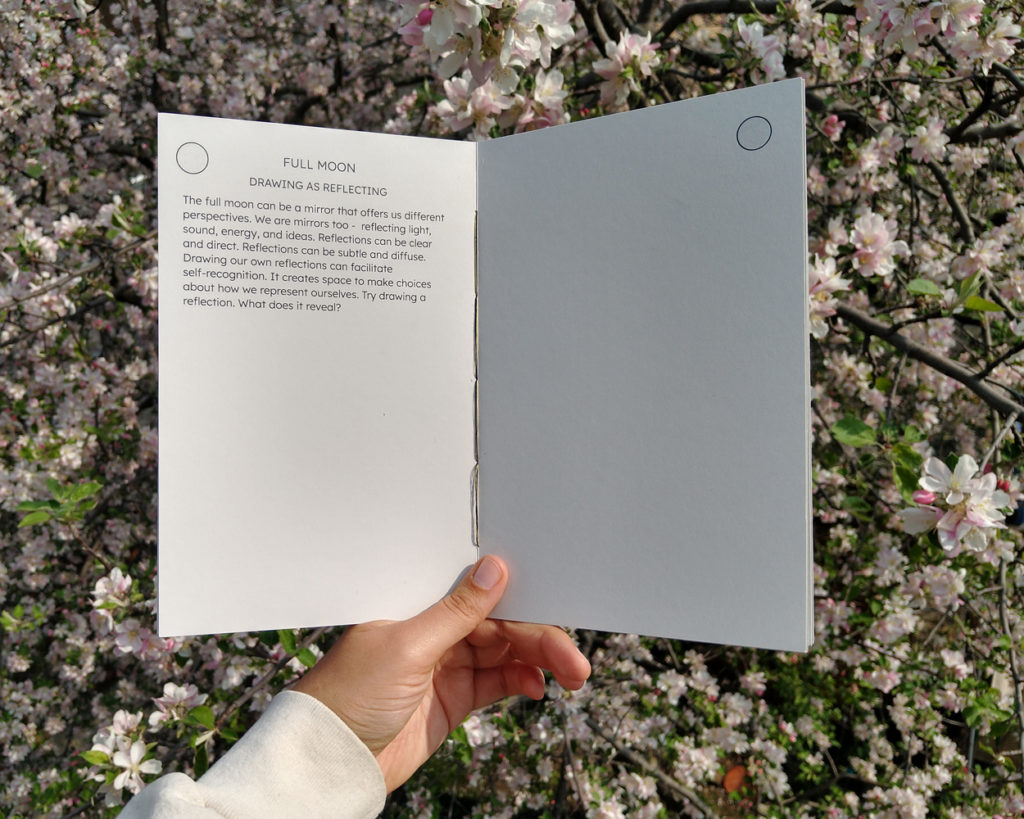 A hand holds a book open above a flowering apple tree. The open page reads: "Full Moon. Drawing as Reflecing. The full moon can be a mirror that offers us different perspectives. We are mirrors too - reflecting light, sound, energy, and ideas. Reflections can be clear and dirrect. Reflections can be subtle and diffuse. Drawing our own reflections can facilitate self-recognition. It creates space to make choices about how we represent ourselves. Try drawing a reflection. What does it reveal?"