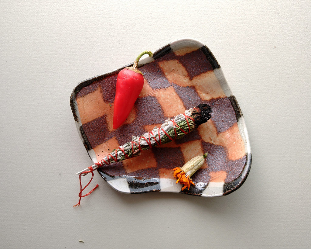 A small square ceramic dish with a nerikomi pattern of contrasting squares hold a hot pepper, a dried flower, and a bundle of herbs for burning.