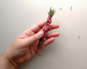 A small bundle of dried rosemary branches and rose petals bound together with a thick, smooth thread.