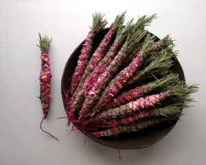 Small bundles made of rosemary branches and herbs petals are stacked in a shallow ceramic bowl.