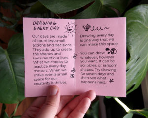 Holding the booklet open to a small essay called "Drawing Every Day". There are illustrations including a talking sun and a tumbling butterfly.