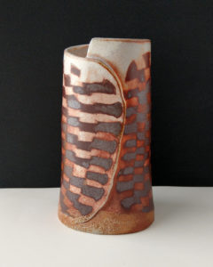 A large ceramic vase sits on the table. The vases surface is covered in a contrasting pattern of smooth, pale clay and rough, dark clay.