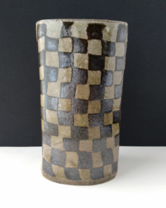 A large ceramic vase sits on the table. The vases surface is covered in a contrasting pattern of smooth, pale clay and rough, dark clay.