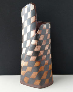 A large ceramic vase sits on the table. The vases surface is covered in a contrasting pattern of smooth, pale clay and rough, dark clay.