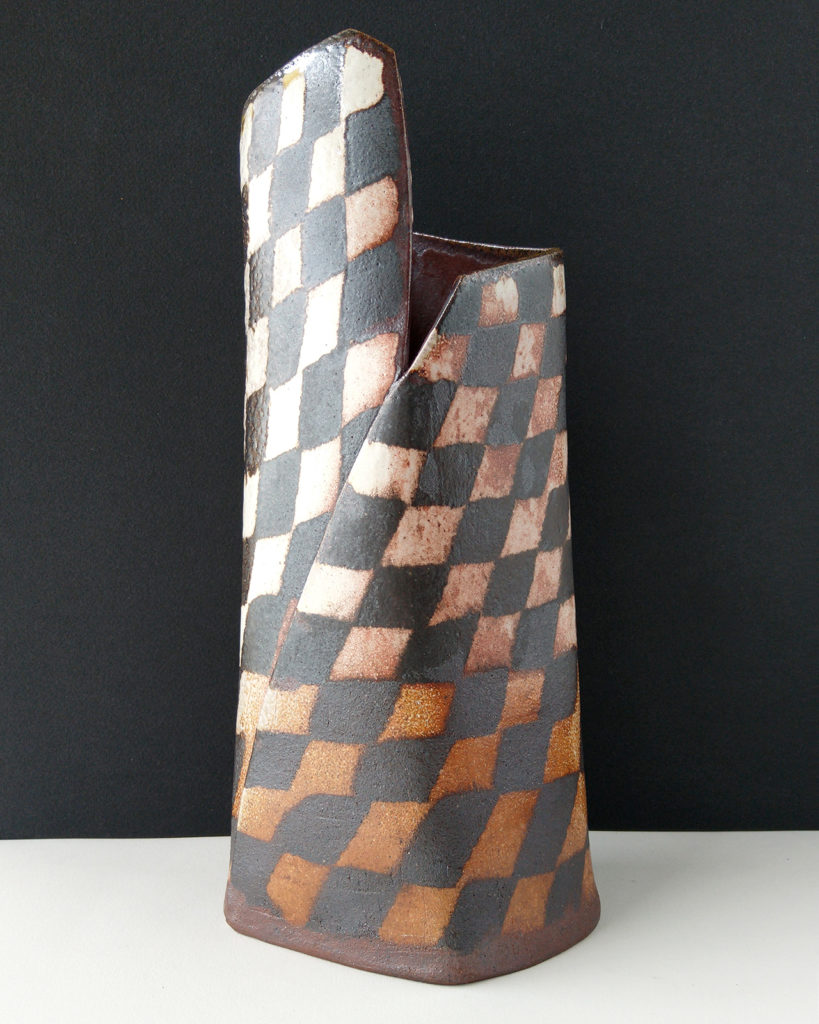 A large ceramic vase sits on the table. The vases surface is covered in a contrasting pattern of smooth, pale clay and rough, dark clay.