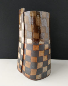 A large ceramic vase sits on the table. The vases surface is covered in a contrasting pattern of smooth, pale clay and rough, dark clay.